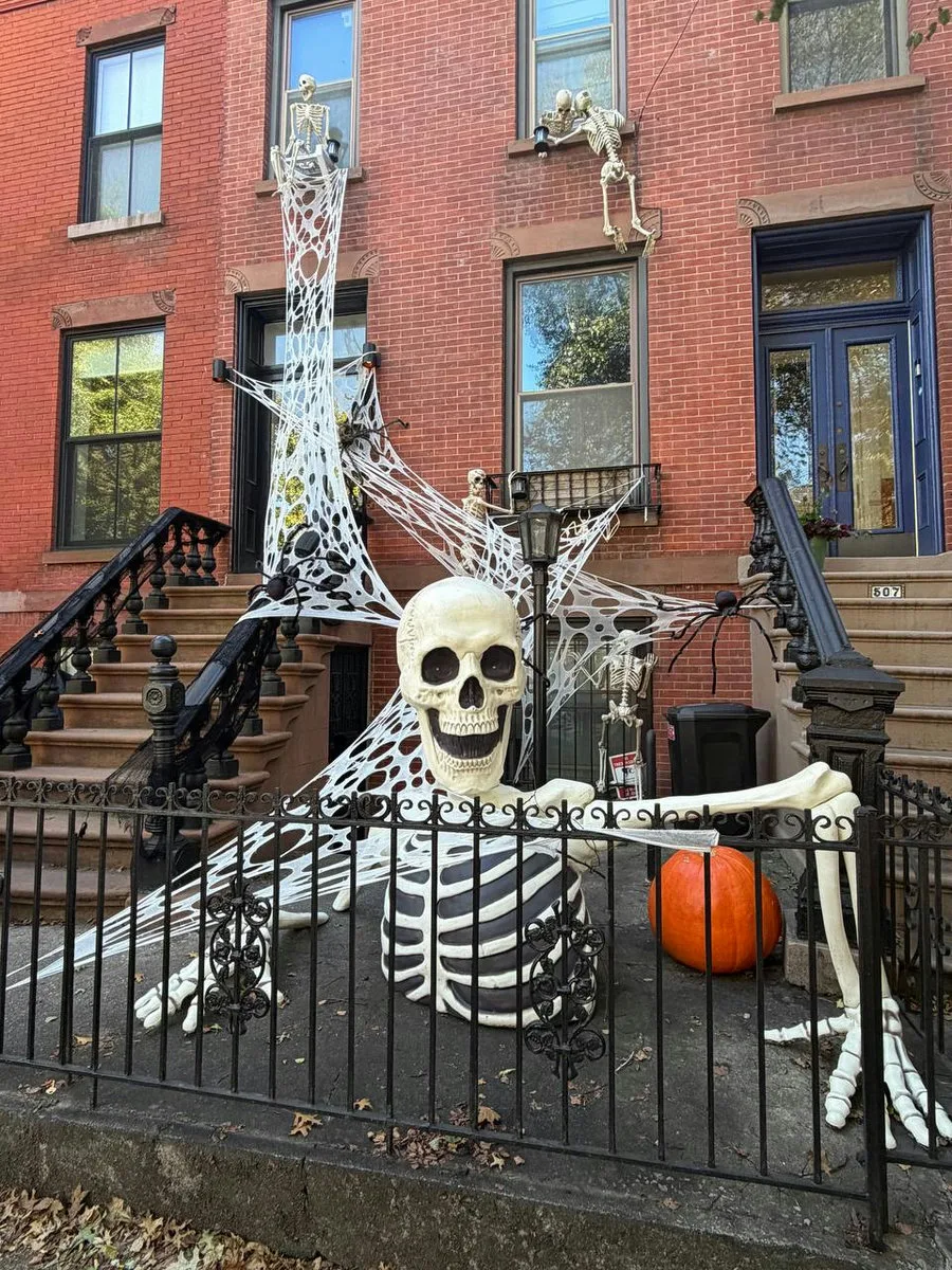A skeleton wrapped in spiderwebs against a brick facade with a pumpkin